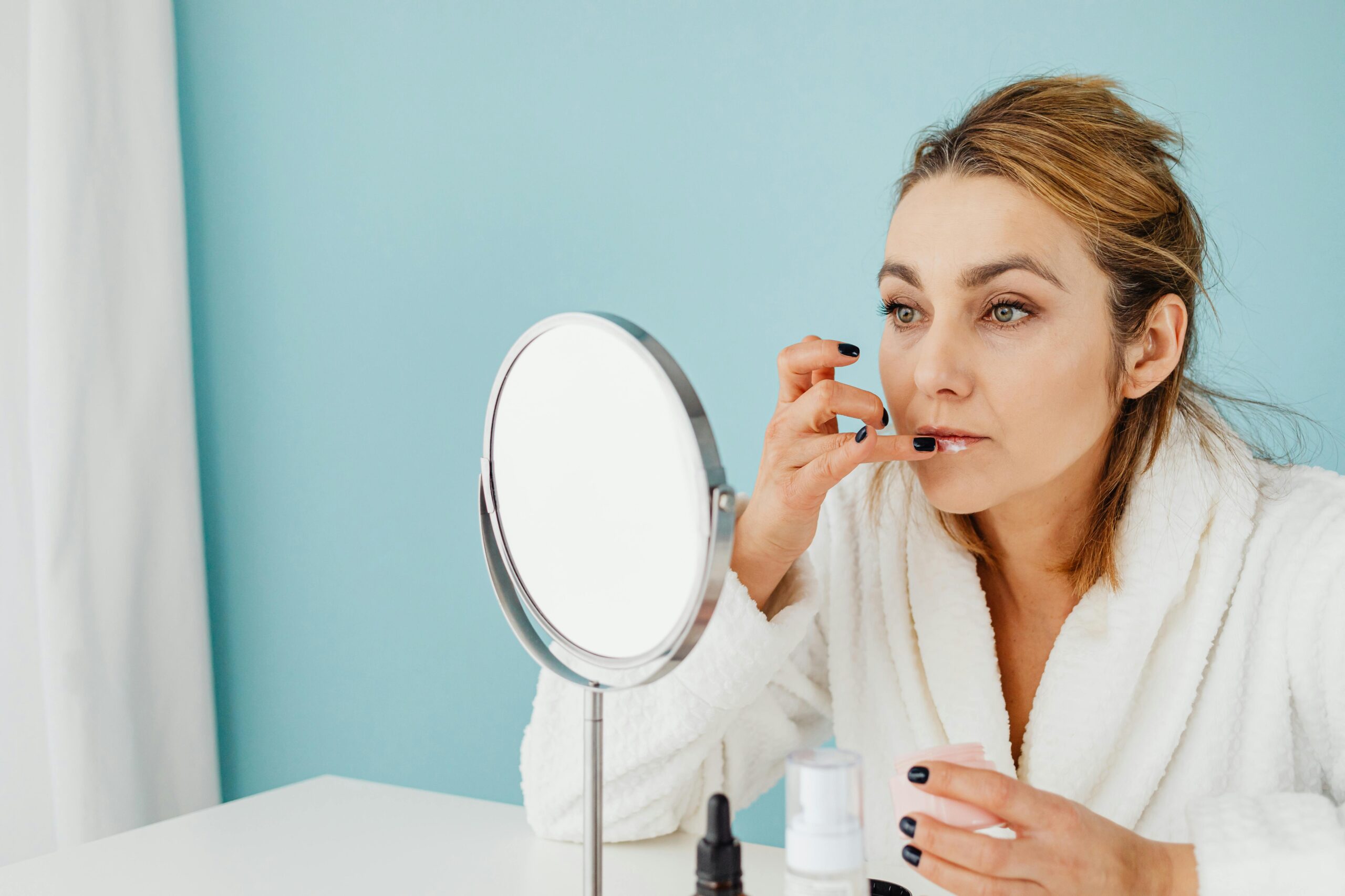 Caucasian woman in a bathrobe applies skincare in front of the mirror against a pastel blue background.