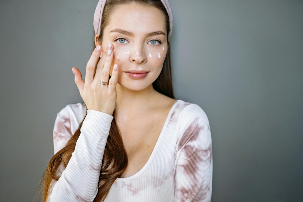 A young woman applies skincare cream to her face, promoting beauty and skincare routine.